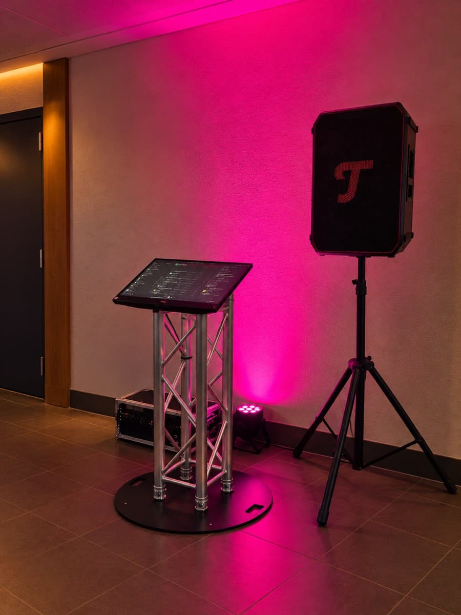 Musiktheke mit Touchscreen auf Traversenständer neben einer Lautsprechersäule, die Wand in pinkes Licht getaucht